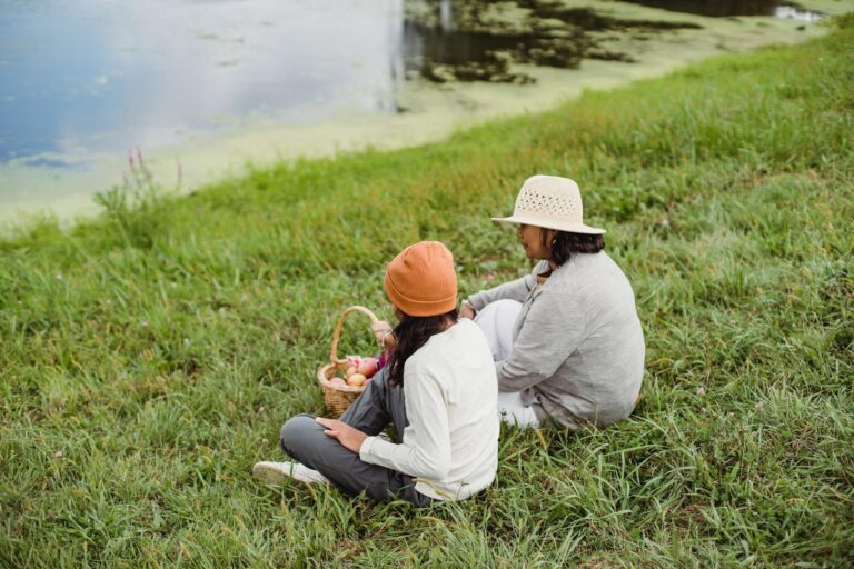 Daughter and mother sitting on grassy shore with basket of apples