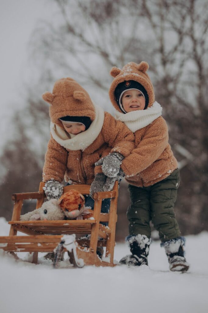 Adorable kids in bear-themed jackets playing in the snow with a wooden sled.